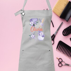 May include: Gray apron with a pocket, featuring a floral design with hair styling tools and the name "Olivia" in orange text.