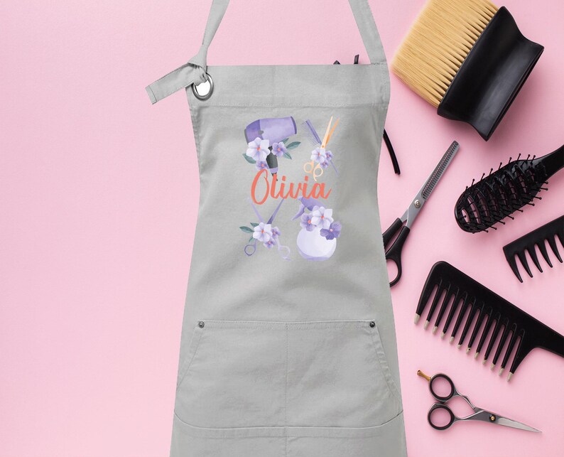 May include: Gray apron with a pocket, featuring a floral design with hair styling tools and the name "Olivia" in orange text.