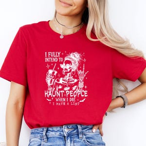 May include: Red t-shirt with a white graphic of a skeleton wearing sunglasses, holding a drink, and giving a rock-on hand gesture. The text reads "I FULLY INTEND TO HAUNT PEOPLE WHEN I DIE I HAVE A LIST."