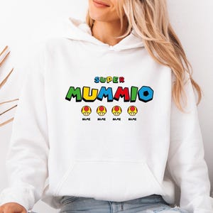 May include: A white hooded sweatshirt featuring a graphic print on the chest. The text reads "SUPER MUMMIO" in colourful, blocky letters. Below the text are four yellow and red mushroom icons, each with the word "NAME" printed underneath it.