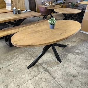 Può includere: Tavolo ovale in legno con base in metallo nero. Il tavolo ha una finitura in legno naturale ed è sostenuto da quattro gambe che formano una X.