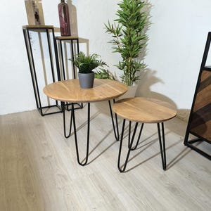 Puede incluir: Dos mesas auxiliares redondas de madera con patas de horquilla negras. La mesa más grande está en primer plano y la más pequeña está detrás. Ambas mesas tienen un acabado de madera natural.
