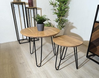 May include: Two round wooden side tables with black hairpin legs. The larger table is in the foreground and the smaller table is behind it. Both tables have a natural wood finish.