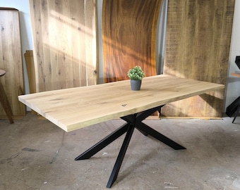 May include: A large wooden dining table with a live edge top and a black metal X-shaped base. The table is made of light wood and has a natural finish. The base is made of sturdy metal and has a black powder coat finish.