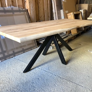 Könnte beinhalten: Ein Holztisch mit einer Live-Edge-Platte und einem schwarzen Metall-Dreibein-Fuß. Der Tisch steht in einer Lagerhalle.