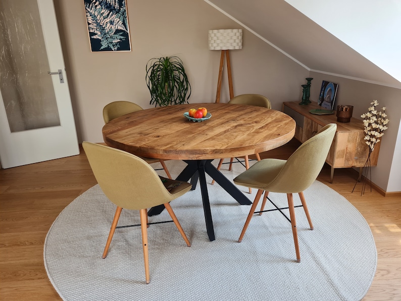 May include: A round wooden dining table with a black metal base and four light green chairs. The table is set with a bowl of fruit and a white rug is under the table.