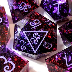 Puede incluir: Un conjunto de dados de color morado y rojo con números blancos y purpurina incrustada en la resina. Los dados son de varias formas, incluyendo un d20, d12, d10, d8, d6 y d4.