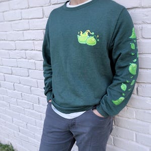 May include: A dark green sweatshirt with a white crew neck collar. The sweatshirt features a pixelated green design on the chest and down the left sleeve. The design includes two green creatures with yellow accents and green leaf shapes.
