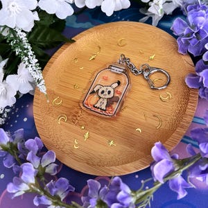 Puede incluir: Un llavero acr&iacute;lico transparente con un personaje de fantasma de dibujos animados dentro de un tarro, con una calabaza y hojas de oto&ntilde;o. El llavero tiene una cadena y un cierre plateados. El llavero est&aacute; sobre un plato de madera con detalles de estrellas y lunas doradas, rodeado de flores blancas y moradas.