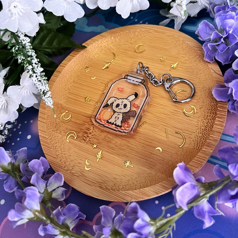 Puede incluir: Un llavero acr&iacute;lico transparente con un personaje de fantasma de dibujos animados dentro de un tarro, con una calabaza y hojas de oto&ntilde;o. El llavero tiene una cadena y un cierre plateados. El llavero est&aacute; sobre un plato de madera con detalles de estrellas y lunas doradas, rodeado de flores blancas y moradas.