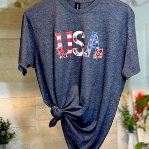 May include: A heathered dark gray short-sleeve t-shirt featuring the text USA printed across the chest. The letters are decorated with red and white stripes, blue with white stars, and floral accents in shades of red, pink, and green.