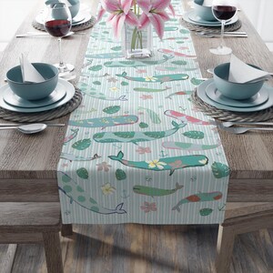 Op de afbeelding: Lichtblauwe tafelloper met een herhalend patroon van walvissen, bloemen en bladeren. De walvissen zijn in verschillende tinten blauw, groen en roze, met bloemen- en polkadotontwerpen. De achtergrond heeft verticale witte strepen. De loper ligt op een houten tafel.