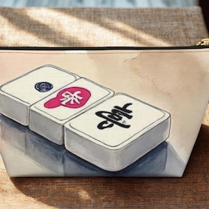 May include: A zipped pouch featuring a printed illustration of three white mahjong tiles arranged in a row. The tiles display distinct black and pink symbols on their surfaces. The pouch has a light-coloured background with soft watercolour-style shading.