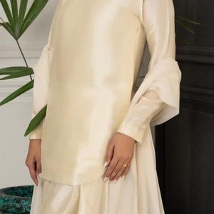 May include: A cream-colored, long-sleeved dress with a flowing skirt and a keyhole neckline. The dress is made of a silky fabric and has a subtle sheen.