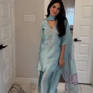 Puede incluir: Conjunto salwar kameez azul claro. La parte superior presenta un adorno en forma de lazo con detalles de cuentas. El conjunto incluye pantalones a juego y un dupatta transparente. Se usa con sandalias de punta abierta.