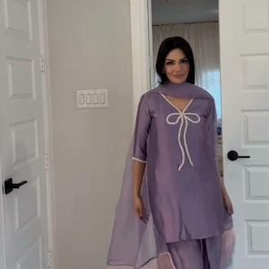 Puede incluir: Un conjunto salwar kameez de color lila. La parte superior presenta un dise&ntilde;o de lazo con ribete blanco. El atuendo incluye pantalones a juego y una dupatta transparente con bloques de color pastel. El conjunto se usa con sandalias.