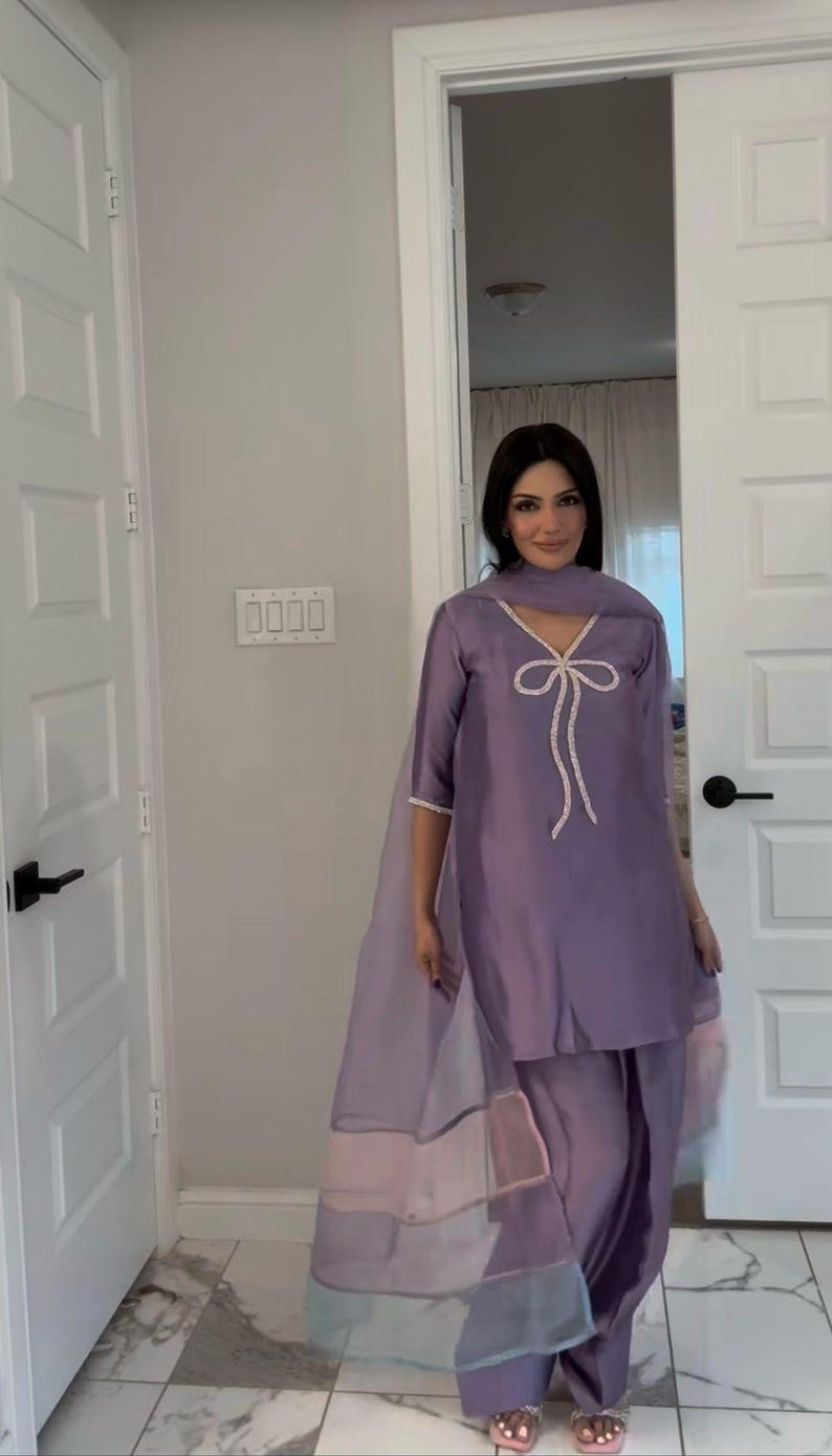 Puede incluir: Un conjunto salwar kameez de color lila. La parte superior presenta un dise&ntilde;o de lazo con ribete blanco. El atuendo incluye pantalones a juego y una dupatta transparente con bloques de color pastel. El conjunto se usa con sandalias.