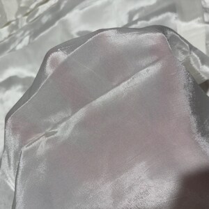 May include: A close-up of white sheer fabric with a smooth, silky texture. The fabric is folded and draped, creating soft folds and wrinkles.