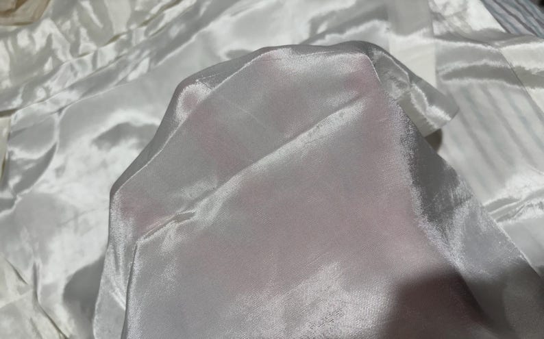 May include: A close-up of white sheer fabric with a smooth, silky texture. The fabric is folded and draped, creating soft folds and wrinkles.
