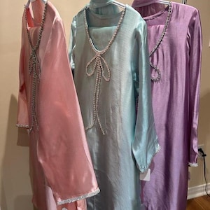Puede incluir: Tres vestidos de seda en colores pastel cuelgan de una percha. Los vestidos son rosa claro, verde agua y lavanda. Cada vestido tiene un escote y un ribete enjoyados. Los vestidos son de manga larga y tienen un dise&ntilde;o fluido.