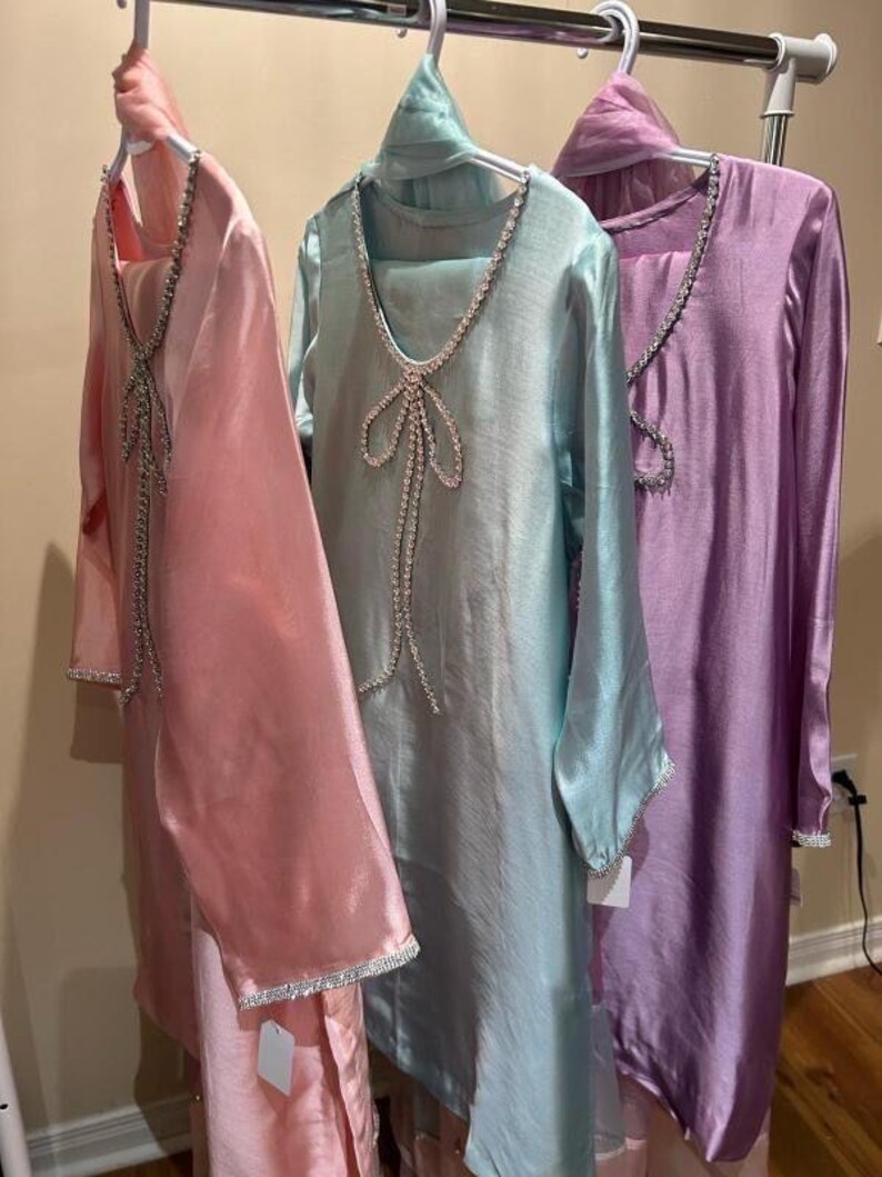 Puede incluir: Tres vestidos de seda en colores pastel cuelgan de una percha. Los vestidos son rosa claro, verde agua y lavanda. Cada vestido tiene un escote y un ribete enjoyados. Los vestidos son de manga larga y tienen un dise&ntilde;o fluido.
