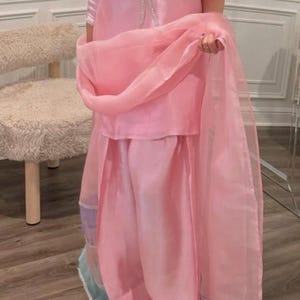 Puede incluir: Un conjunto de dos piezas rosa claro con una t&uacute;nica con cuello en V y pantalones de pernera ancha. El conjunto incluye una bufanda transparente a juego. La t&uacute;nica tiene un lazo decorativo y los pantalones un ribete decorativo.