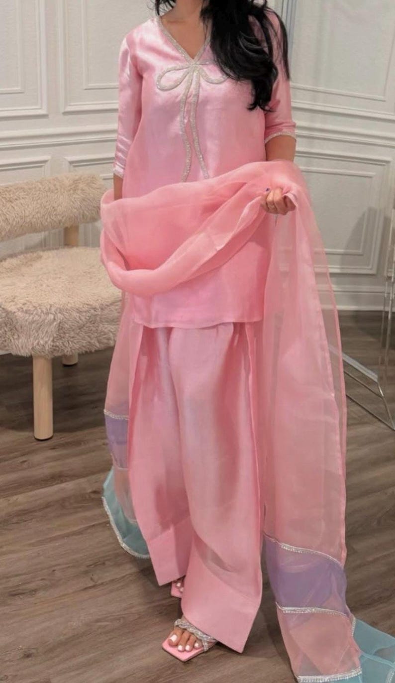 Puede incluir: Un conjunto de dos piezas rosa claro con una t&uacute;nica con cuello en V y pantalones de pernera ancha. El conjunto incluye una bufanda transparente a juego. La t&uacute;nica tiene un lazo decorativo y los pantalones un ribete decorativo.