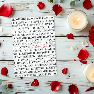 May include: Romantic still life with a white wooden background. A card with "I Love You" text is surrounded by red rose petals, a red rose, and two lit candles in glass holders. The scene evokes love and affection.