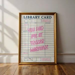 Puede incluir: Impresión enmarcada con un diseño de tarjeta de biblioteca vintage. La tarjeta tiene líneas de cuadrícula azules y texto rosa que dice "YOU HAD ME AT TRIGGER WARNINGS". El marco es dorado.