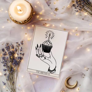 Może przedstawiać: Czarno-biała ilustracja dłoni trzymającej babeczkę ze świeczką, z napisem "THE BIRTHDAY". Obrazek znajduje się na białej karcie, otoczony zapaloną świecą, lawendą i złotymi gwiazdami na białej tkaninie.