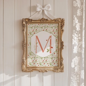 Op de afbeelding: Een ingelijste print met de letter "M" in koraal, versierd met bloemen- en aardbeimotieven. De gouden sierlijke lijst wordt opgehangen door een wit lint en strik. De print is ontworpen als decoratieve wanddecoratie.
