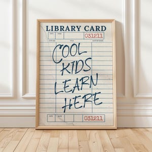 May include: A framed print featuring a vintage library card design on off-white paper with blue horizontal lines and vertical columns. The text reads LIBRARY CARD at the top, 031211 in red, and COOL KIDS LEARN HERE in large dark blue script.