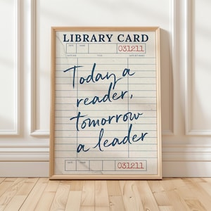 May include: A framed print featuring a vintage library card design. The print has a cream-colored background with blue lines and text. The text reads "Today a reader, tomorrow a leader" in a handwritten style. The frame is a light brown color.
