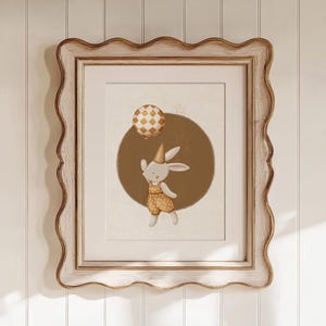 Puede incluir: Una impresión enmarcada de un lindo conejito con un gorro de fiesta y sosteniendo un globo. El conejito lleva un atuendo de lunares amarillos y marrones y está de pie sobre un círculo marrón. El fondo es blanco.