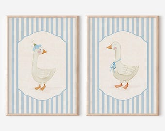May include: Two framed prints featuring watercolor illustrations of geese. One goose wears a blue flower hat, the other a blue bow. The background is a light blue and white striped pattern, with a cream-colored border around the geese.