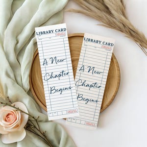 Könnte beinhalten: Zwei rechteckige Lesezeichen aus Papier, die wie alte Bibliothekskarten gestaltet sind. Jedes zeigt blaue horizontale Linien, den Text "LIBRARY CARD" oben und den kursiven Schriftzug "A New Chapter Begins" in der Mitte mit roten Stempeln.