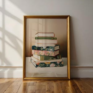 May include: A framed art print featuring a stack of books. The books have various covers, including floral patterns and solid colors. The frame is gold, and the artwork is displayed against a neutral background.