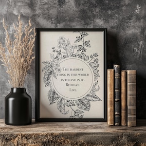 May include: A framed art print with botanical illustrations and the text "The hardest thing in this world is to live in it. Be brave. Live." A black vase with dried plants and vintage books are also in the image.