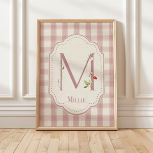 Op de afbeelding: Een ingelijste print met een roze en witte ruitjespatroon. De centrale designelement is een roze letter "M" versierd met bloemmotieven. De naam "Millie" staat eronder in een bijpassend roze lettertype.