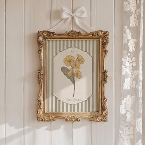 May include: A decorative framed botanical print with yellow flowers and green leaves, set against a striped background. The ornate gold frame is suspended by a white ribbon tied in a bow, creating a vintage aesthetic.