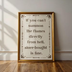 May include: A framed sign with the text "If you can't summon the flames directly from hell, store-bought is fine." The sign has a gold frame and is displayed against a white wall and wooden floor.