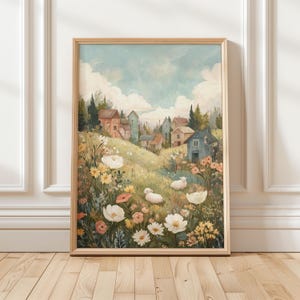 May include: A framed art print showing a rural landscape. The image features a village on a hillside, with houses in various colours, sheep, and wildflowers in white, yellow, and pink. The sky is a pale blue with clouds. The frame is made of light wood.