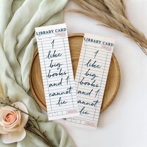 Peut inclure: Deux marque-pages en forme de cartes de bibliothèque avec l'inscription manuscrite "I like big books and I cannot lie". Ils sont posés sur une assiette en bois, avec un tissu vert clair et des herbes séchées en arrière-plan.