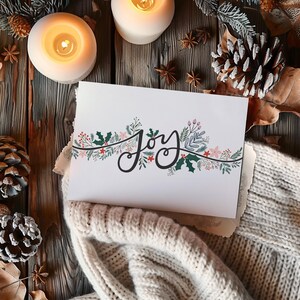 Könnte beinhalten: Eine weiße Karte mit dem Wort "Joy" in eleganter Schrift, verziert mit einem floralen Rand aus roten Beeren, rosa Blüten und grünen Blättern. Die Karte liegt auf einer cremefarbenen Strickoberfläche, mit brennenden Kerzen und Tannenzapfen im Hintergrund.