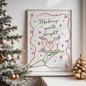 Op de afbeelding: Een ingelijste print met de tekst "Making spirits bright" in groen schrift, met illustraties van cocktails op een dienblad. Het kunstwerk is wit omlijst en geaccentueerd met rode decoratieve elementen. Een kerstboom en een gouden decoratieve boom staan op de voorgrond.