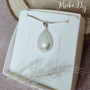 May include: A pear-shaped pendant necklace with the name "Louis" in cursive. The pendant is white with iridescent glitter and is on a silver chain. The necklace is presented in a white box.