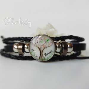 以下が含まれることがあります： カラフルなアクセントが付いた木の模様が特徴の、丸いシルバーカラーのチャームが付いた黒い革のラップブレスレットです。「Jasmin」という名前と「18.10.24」の日付がプリントされています。ブレスレットには、シルバーカラーのビーズと編み革ストラップが付いています。