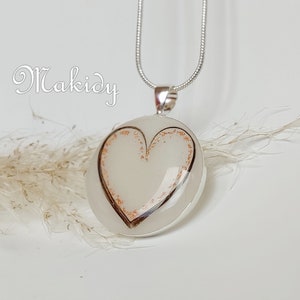 May include: A silver pendant necklace featuring a heart-shaped design. The pendant has a white background with a copper-colored heart outline and copper-colored flecks. The word "Makidy" is in the upper left corner.