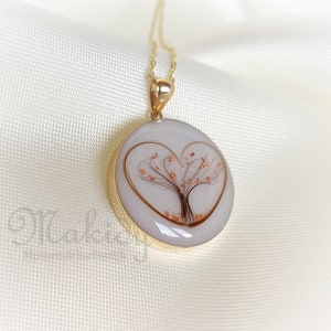 May include: Gold-tone pendant necklace featuring a round, white pendant with a heart-shaped gold outline. Inside the heart is a tree design with brown branches and copper-colored accents. The necklace hangs on a delicate gold chain.