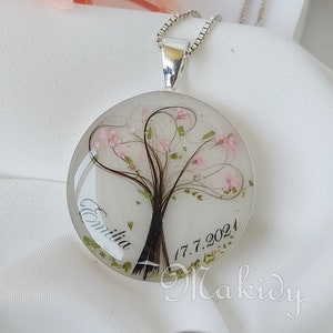 May include: A silver necklace with a round pendant featuring a tree made of hair with pink flowers and green leaves. The pendant is inscribed with the name "Emilia" and the date "17.7.2021".
